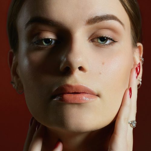 Close-up of a woman's face; she touches her cheek with her fingers, red nails, red background behind her.